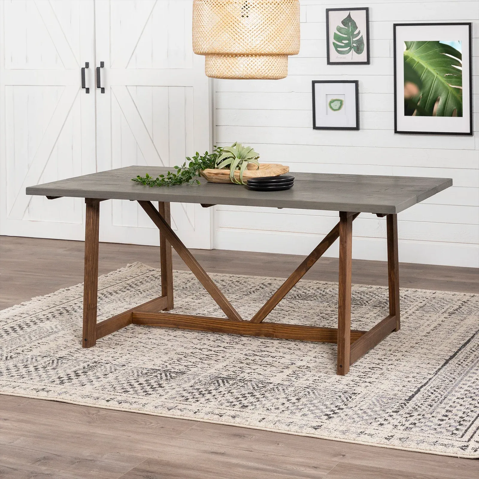 Walker Edison Trestle Dining Table features a concrete top and wooden trestle base on a patterned rug, with simple decor including greenery and black plates. Wall art and a woven light fixture enhance the modern, natural ambiance.