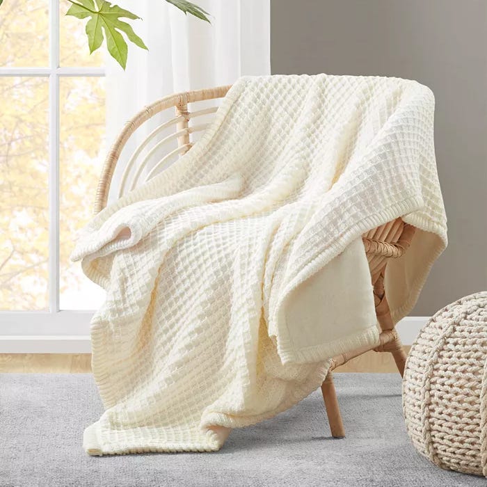 A cream-colored textured throw blanket is draped over a rattan armchair, with a window and a knit pouf nearby.