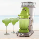 A green Margaritaville drink maker with a clear ice reservoir on top and a blending jar below, next to two margarita glasses filled with a frozen green beverage on a beach backdrop.