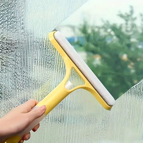 A hand is using a yellow squeegee to clean a soapy glass window.
