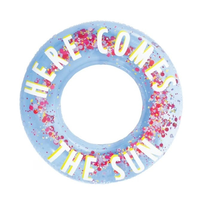 A blue inflatable tube float filled with colorful confetti, featuring the words \“HERE COMES THE SUN\“ in large, white and yellow letters around the center.