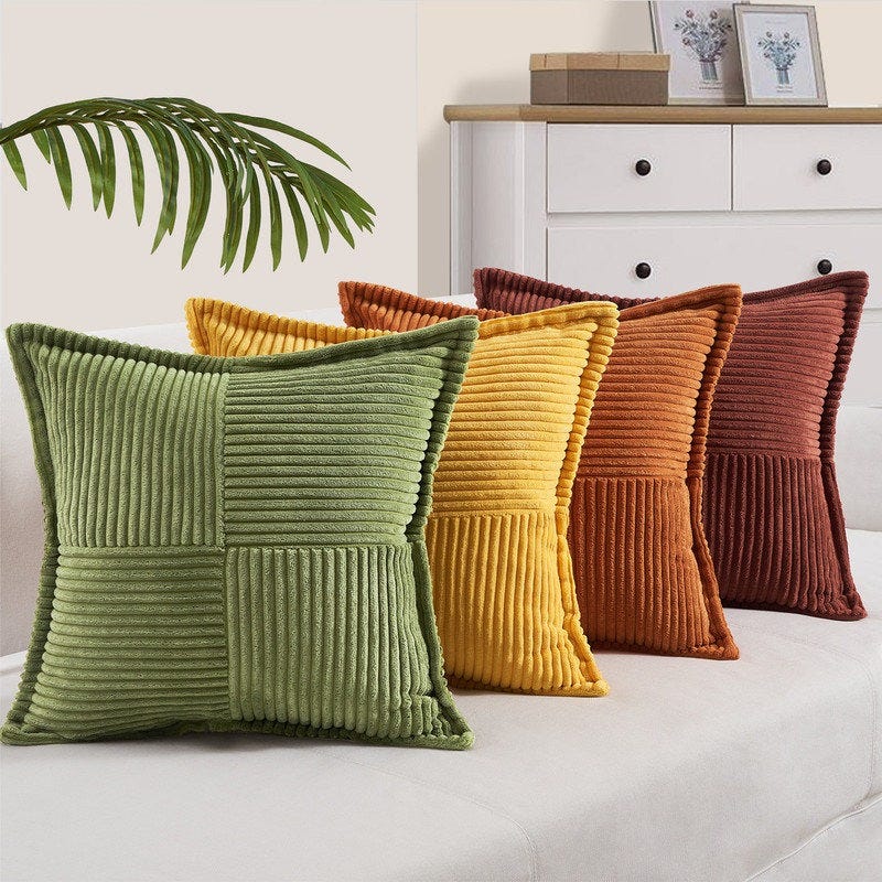 Four decorative corduroy pillows in green, yellow, orange, and brown are arranged on a light-colored sofa. A leaf decor and a dresser with framed pictures are in the background.