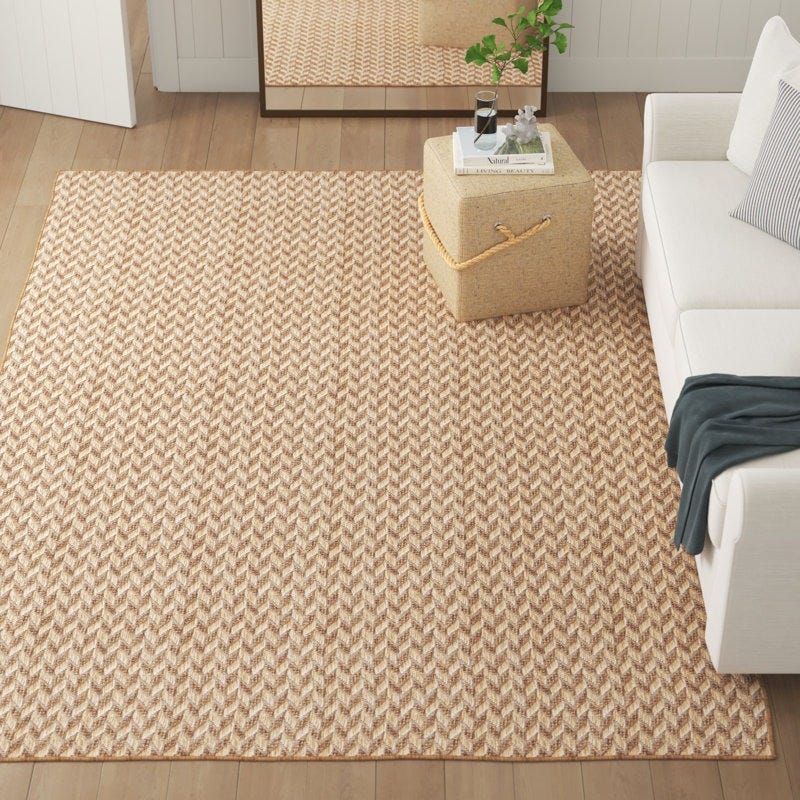 A beige chevron-patterned area rug lies on a wooden floor, next to a white sofa with a striped pillow and a beige pouf with a rope handle holding a book and a plant. A mirror reflecting the rug is partially visible.