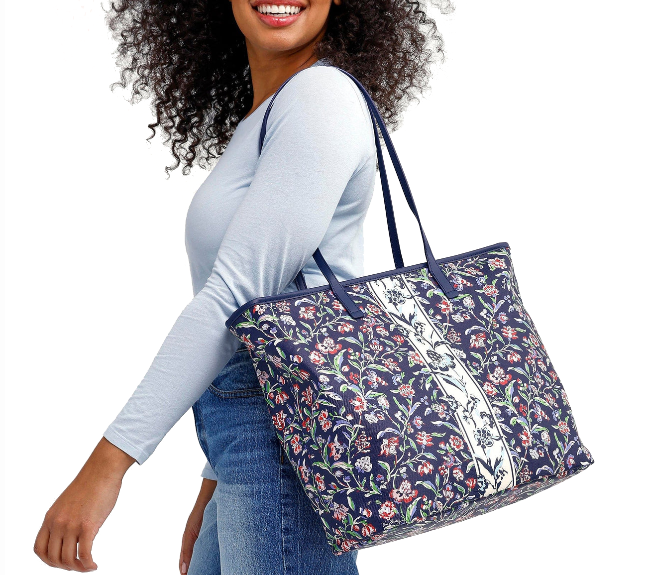 A woman carries a Vera Bradley Large Zip-Top Tote, featuring a navy blue background with colorful floral patterns and a central white stripe. The tote has long shoulder straps, suitable for everyday use.
