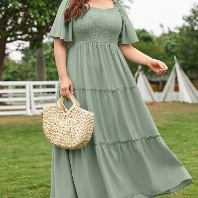 A person is wearing a sage green, short-sleeved, tiered maxi dress and holding a woven straw handbag, outdoors in a grassy area with trees and a fence in the background.