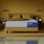 A Casper brand mattress on a bed frame with a brown bedsheet set in a room with a yellow wall and hanging lamps.