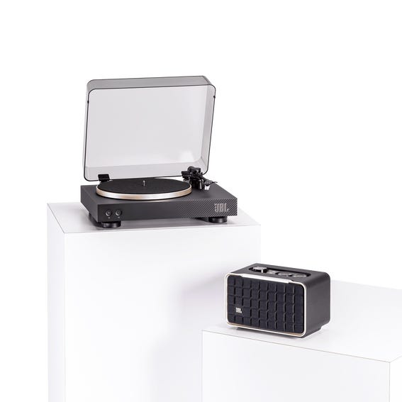 The JBL Music Lover Bundle features a sleek black turntable with a transparent lid and a compact black Bluetooth speaker with a grid design. Both items are placed on white pedestals, highlighting their modern aesthetic.