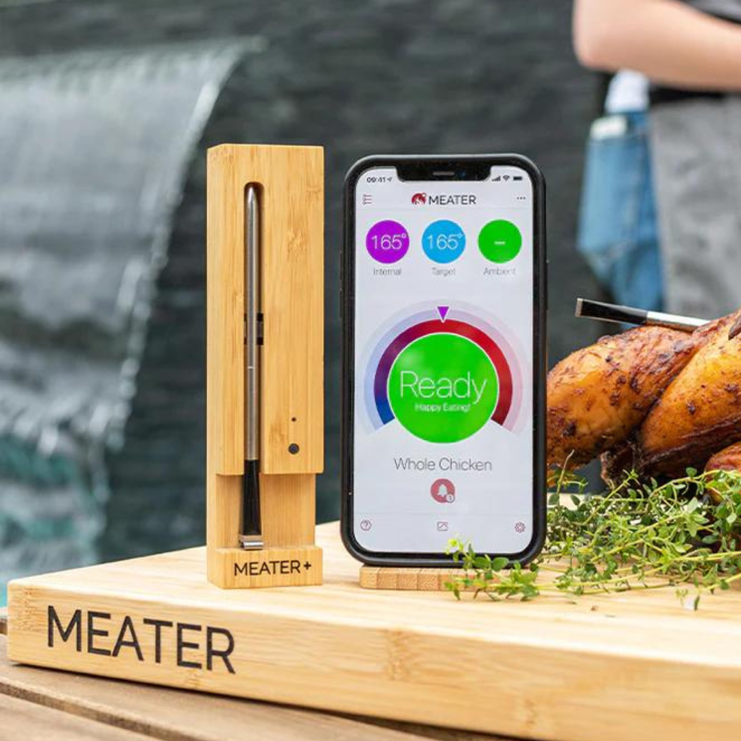 A MEATER+ wireless meat thermometer next to a smartphone displaying cooking temperatures, with a roasted chicken in the background.