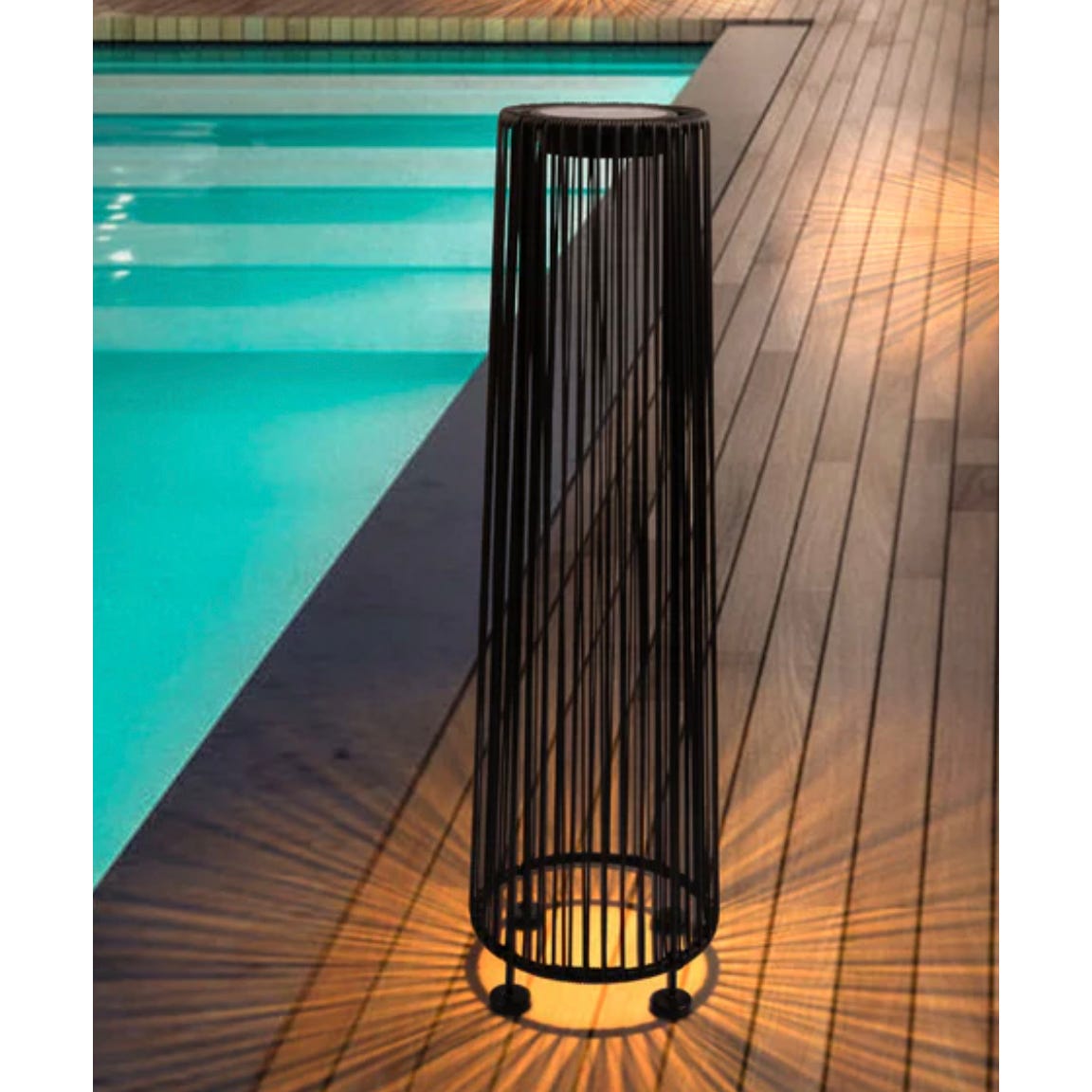 Vertical floor lamp with a cylindrical black metal frame casting a starburst light pattern on a wooden deck next to a swimming pool.