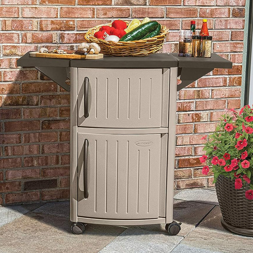 A beige outdoor storage cart with a dark countertop extension, on wheels, beside a brick wall and red flowers. It holds a basket of vegetables and some bottled beverages.