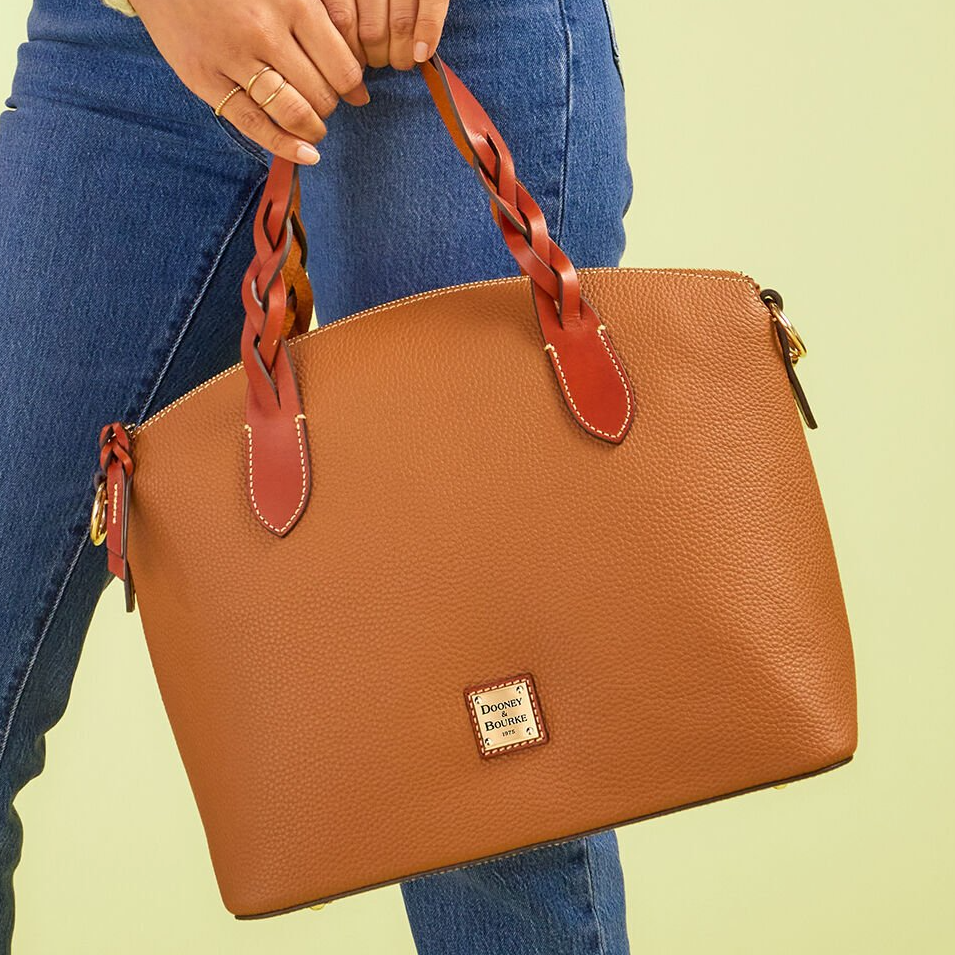 A person holding a tan leather handbag with braided red straps.