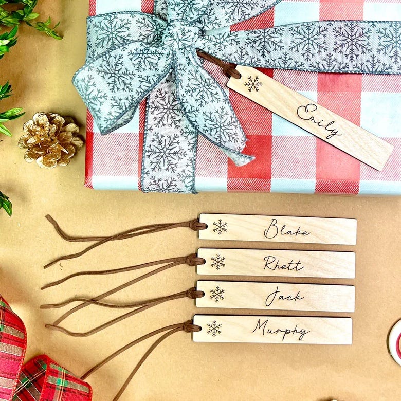 Five custom wooden gift tags with names engraved on them, featuring a small snowflake design and a brown string for attachment.