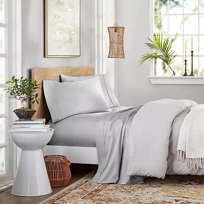 Bed with grey sheets and a light grey throw blanket, accompanied by a white round side table and a wicker pendant lamp.