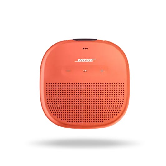 Bose SoundLink Micro Speaker in orange with compact, square design, featuring \“+\“ and \“-\“ buttons on top, speaker grille at the bottom, and water-resistant build. Suitable for portable audio needs.