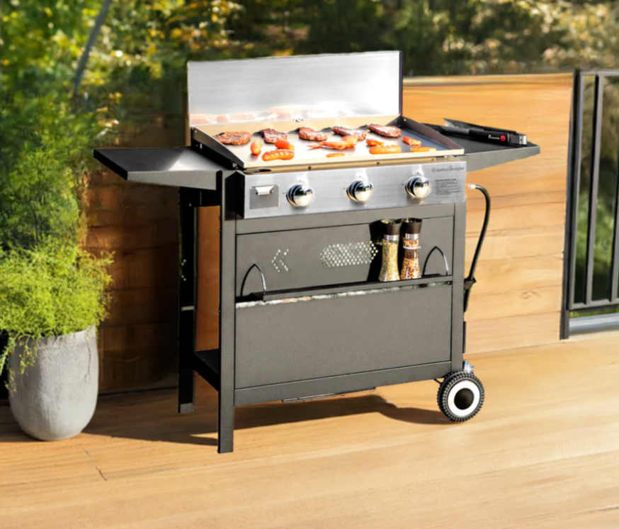 A gas griddle grill featuring three burners with control knobs, side shelves for additional space, and food, including meat and vegetables, cooking on the flat grill surface. A storage area underneath houses condiments and grilling tools, with wheels for mobility.