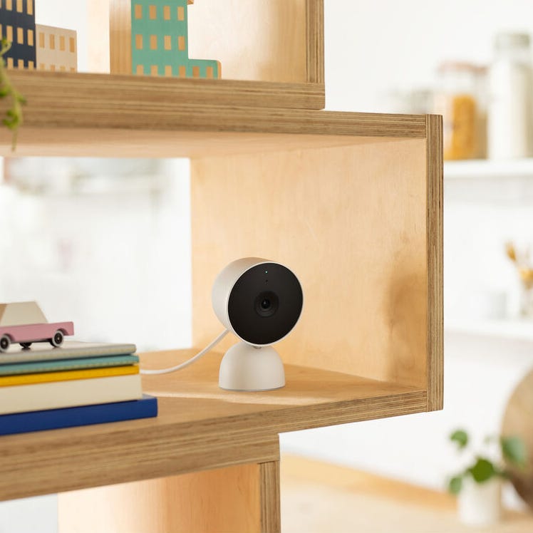 A compact home security camera is displayed on a wooden shelf, alongside decorative items and books.