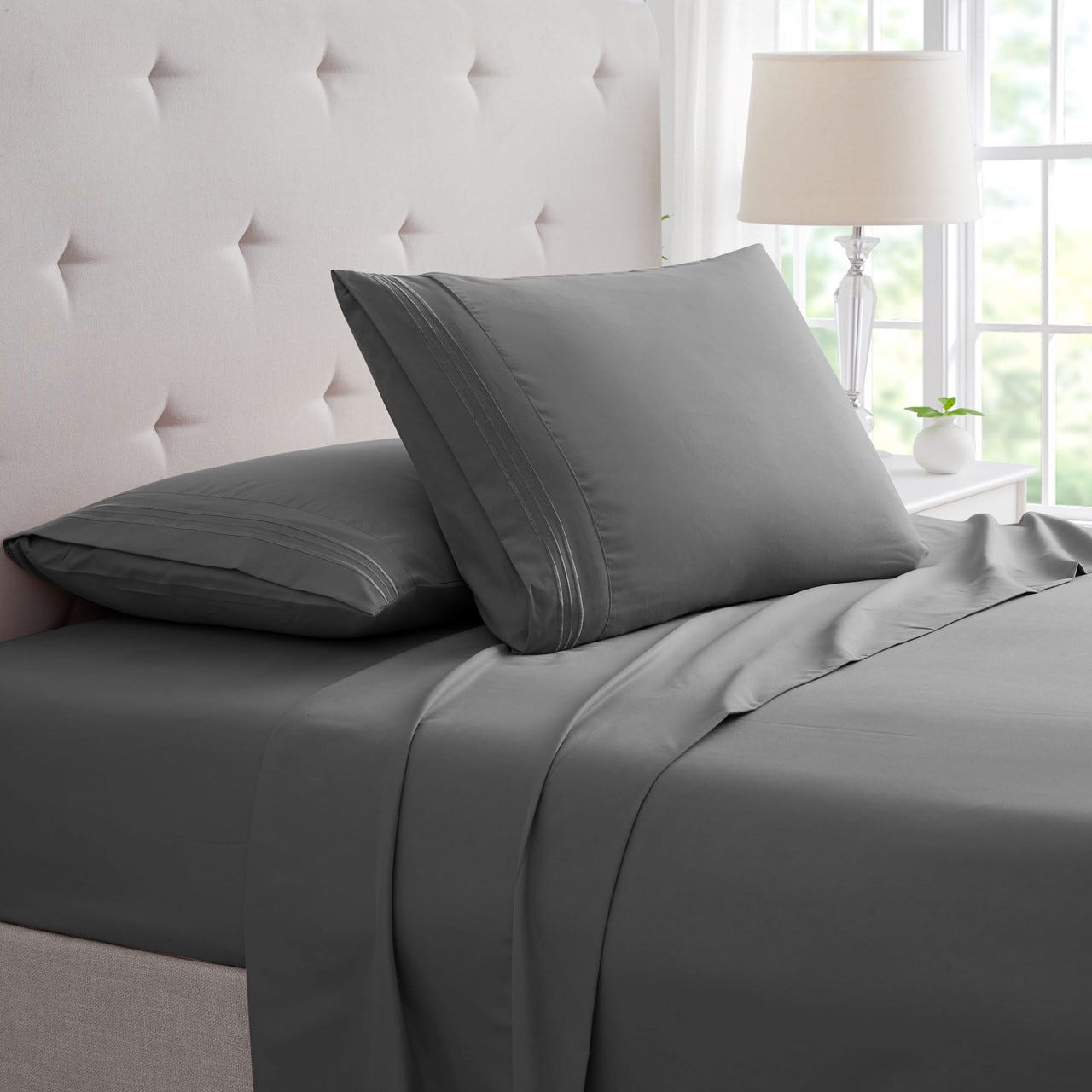Grey bed sheets and pillowcases on a bed, with a tufted headboard and table lamp in the background.