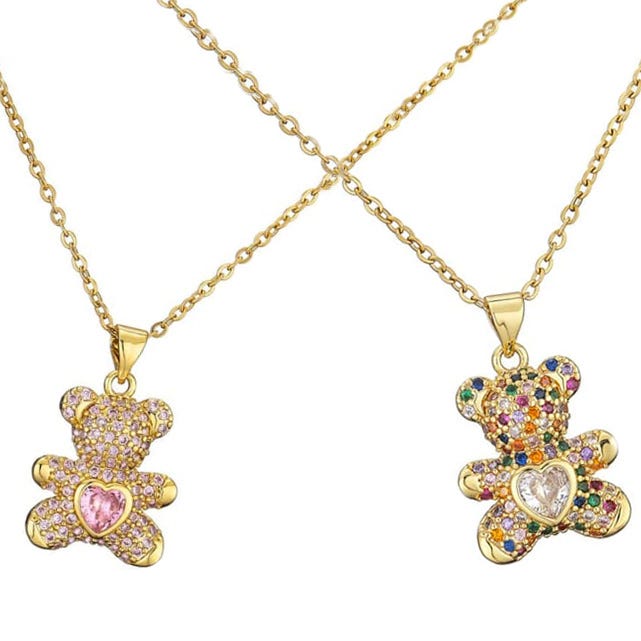 Two gold necklaces featuring teddy bear-shaped pendants adorned with multi-colored crystals. Each bear holds a heart-shaped gem, one pink and the other clear. The chains are thin and gold-toned.