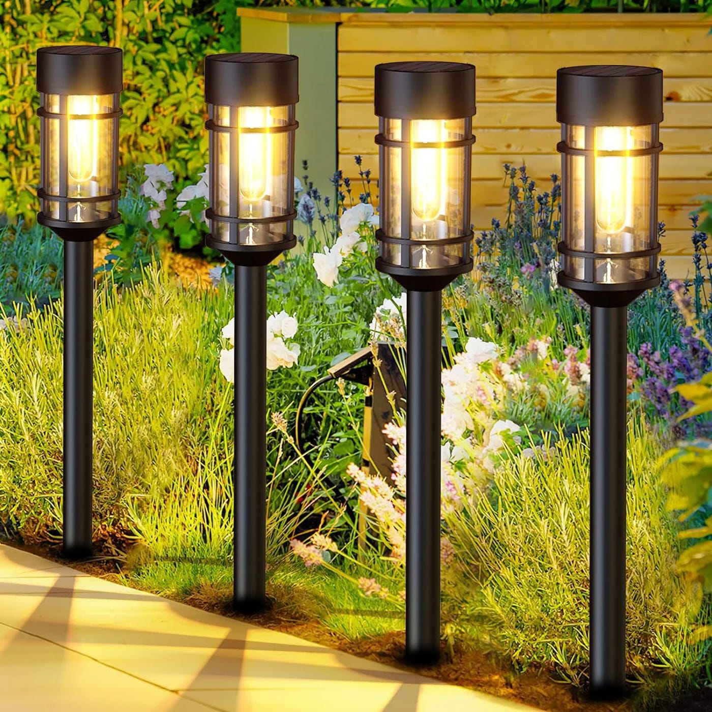 Four garden solar lights on poles illuminating a path with plants in the background.