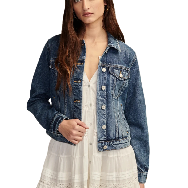 A model wears a blue denim Tomboy Trucker jacket featuring a button front, pointed collar, and flap chest pockets, over a white dress with lace details.