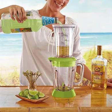 A person is adding ingredients to a Margaritaville Drink Maker, which is positioned next to a bottle of tequila and a plate with lime wedges.