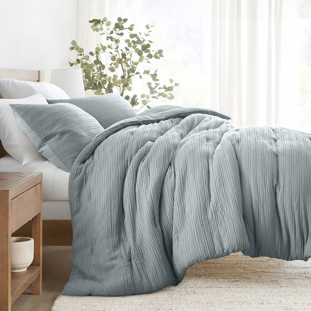 A gray textured comforter on a bed with matching pillows and a white sheet set, next to a wooden nightstand.