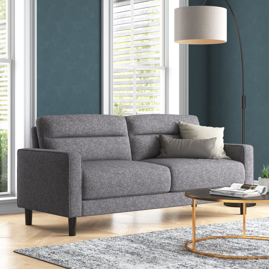 A gray fabric sofa, a round wood and metal coffee table, a floor lamp with a beige shade, and a plush gray rug are shown in a living room setting.