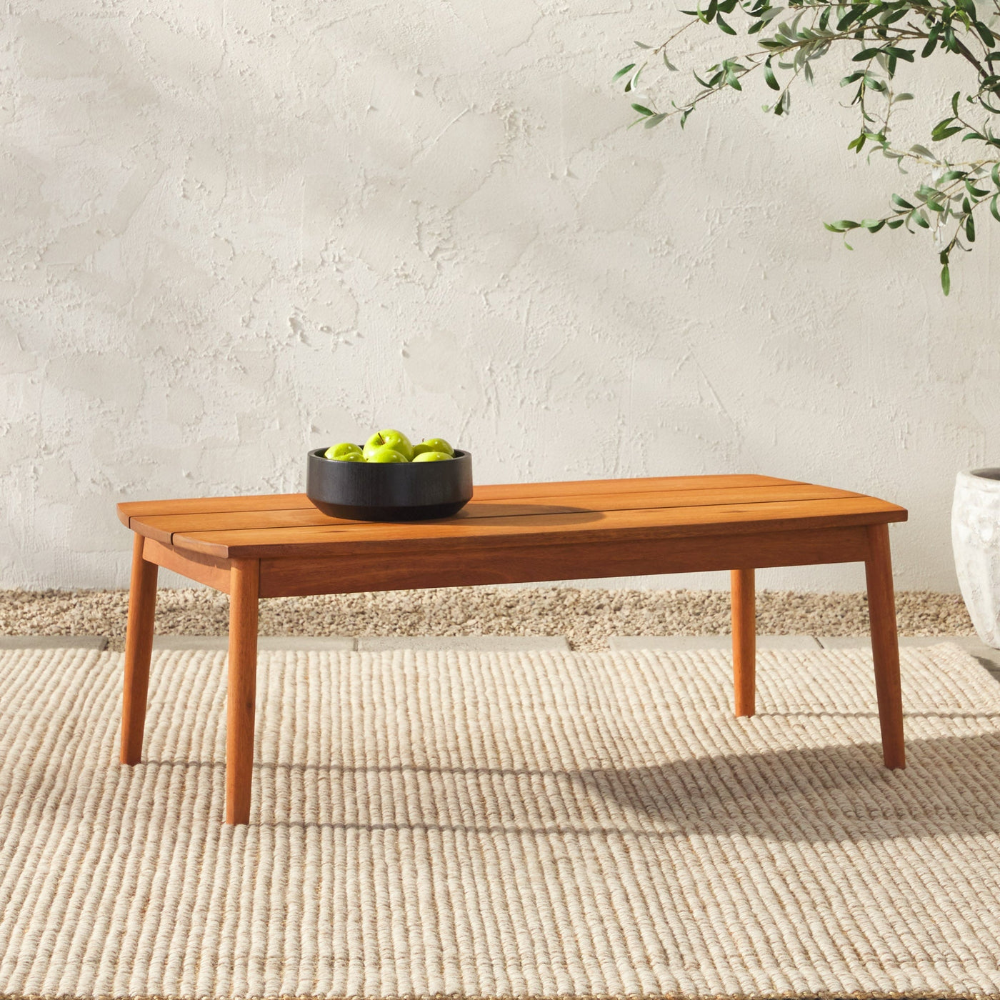 Acacia wood patio coffee table with a natural finish, featuring clean, simple lines and tapered legs. It holds a black bowl filled with green apples and rests on a textured beige rug against a rough-textured wall.