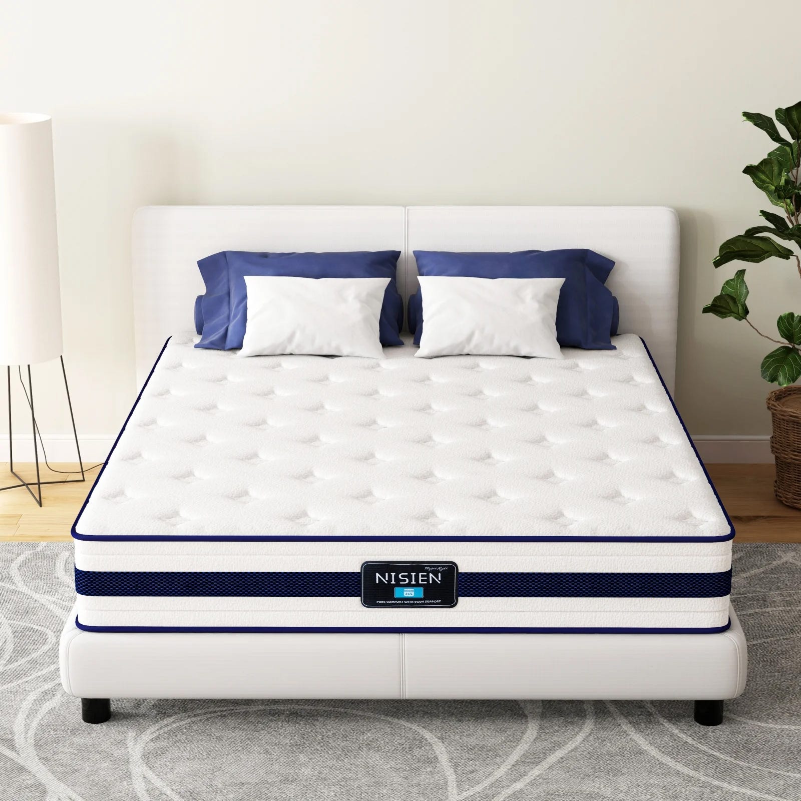 A white, tufted mattress labeled \“NISIEN\“ with blue accents sits on a white bed frame. It is flanked by two blue and white pillows, a plant, and a floor lamp, on a patterned gray rug.