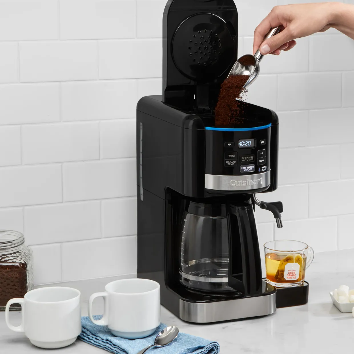 A Cuisinart coffee maker with a digital display, a hand pouring ground coffee, two white cups, and a glass jar of coffee grounds.