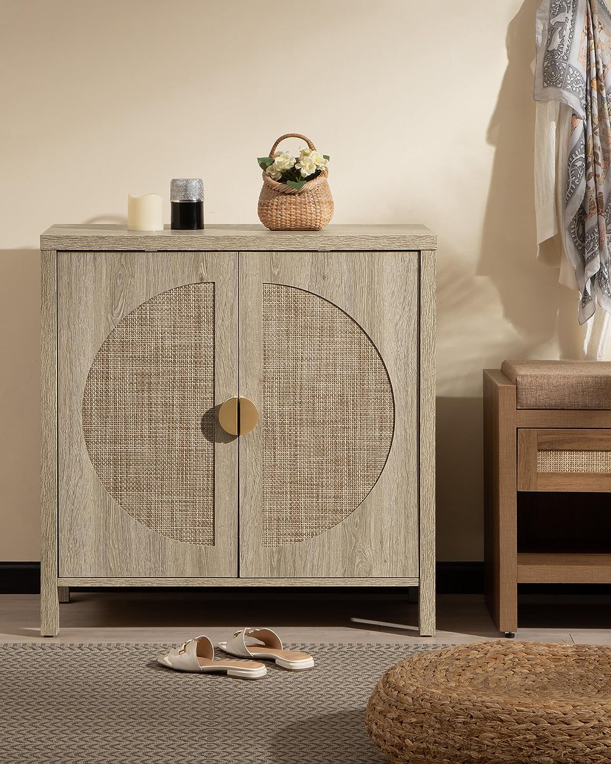 A beige accent storage cabinet features woven rattan door fronts with a circular pattern and a golden round handle. It is topped with a wicker basket with flowers, a candle, and a small black container. Sandals are on a gray-patterned rug next to it.