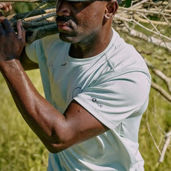 A man wears a light blue Carhartt Sun Defender Tee, featuring short sleeves and a logo on the sleeve, while carrying branches.