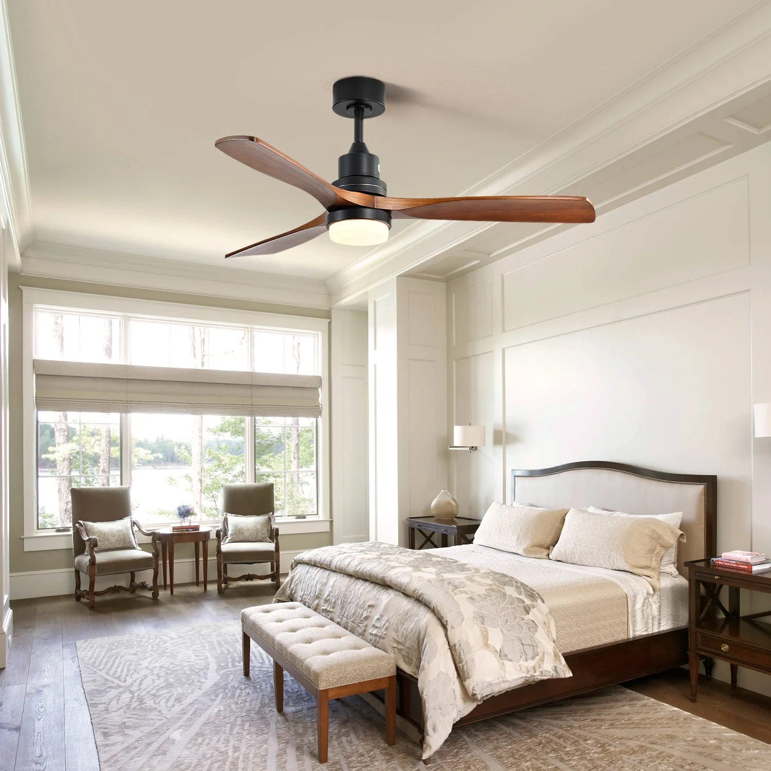 A ceiling fan with light, wooden blades and a dark finish, above a bedroom set with a bed, bench, and two armchairs.