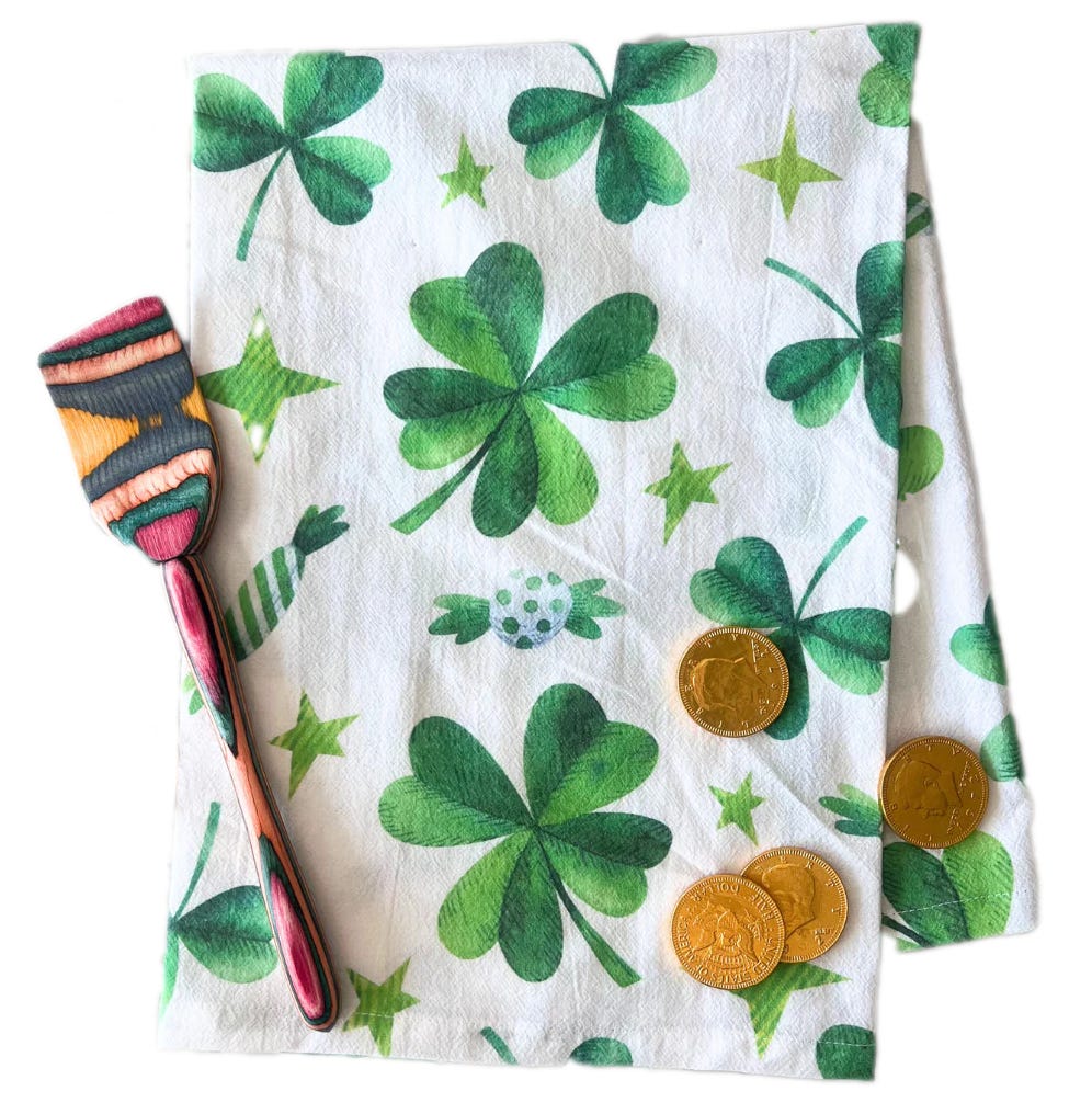White kitchen towel featuring green four-leaf clovers and stars, accompanied by three gold coins and a colorful spatula.