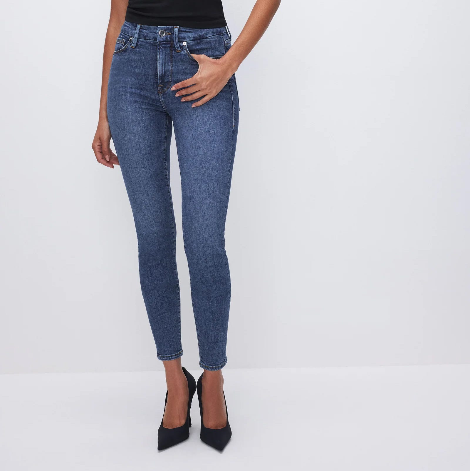 The jeans are a pair of Good American Good Legs Skinny Jeans, featuring a high-rise waist, fitted silhouette through the hips and legs, and a classic blue denim wash. The person is also wearing black high heels.