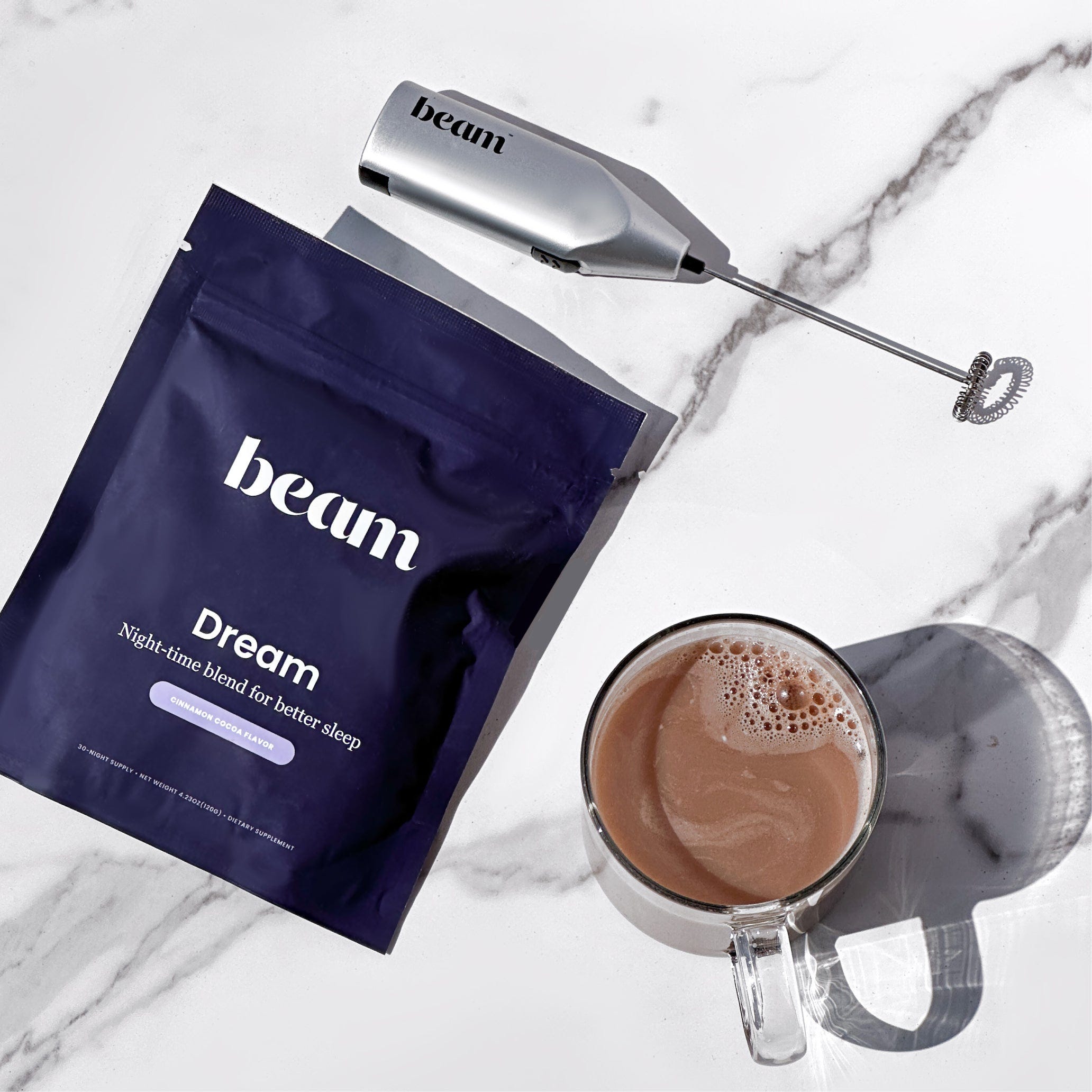 Beam Dream Sleep Powder pouch and a frothy drink in a clear glass mug, with a milk frother lying next to it on a marble surface.
