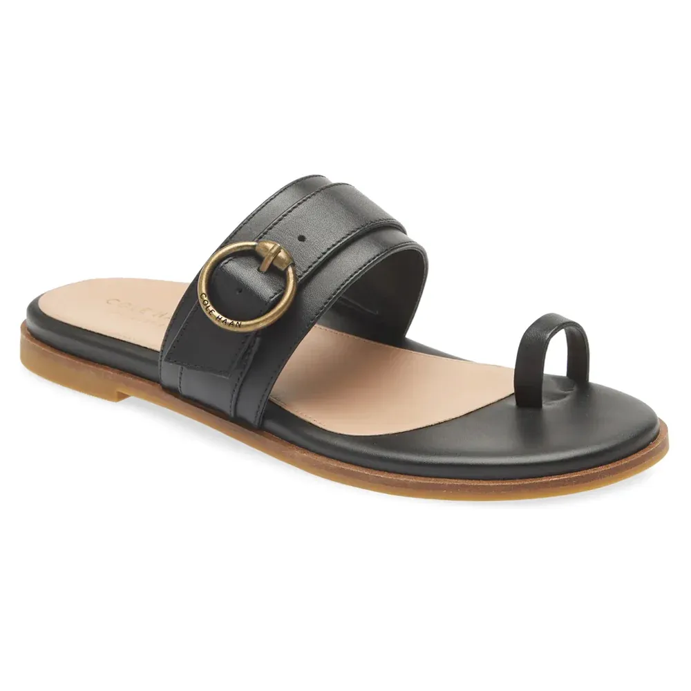 A black flat sandal with a gold-toned circular buckle on the strap.