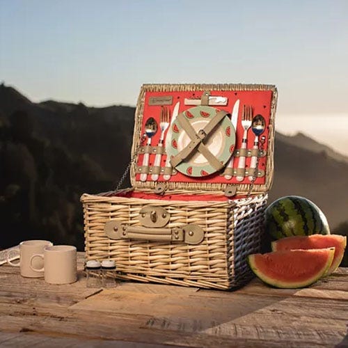 A wicker picnic basket with built-in dining set, a sliced watermelon, and two mugs on a scenic overlook.