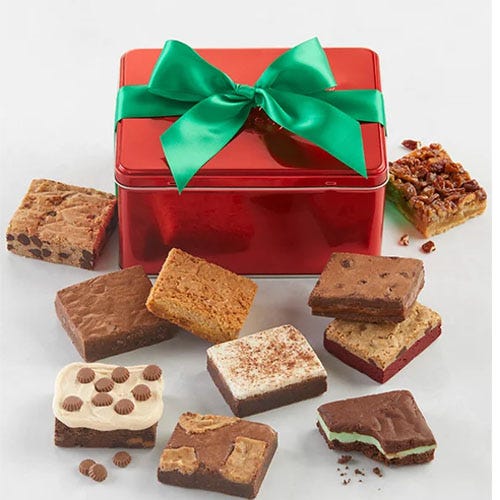 A red tin with a green bow containing an assortment of brownies in various flavors, including those with chocolate chips, nuts, and layered toppings. Some brownies have visible toppings like peanut butter cups and mint layers.