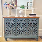 Decorative blue sideboard with ornate cut-out door panels, topped with a vase of artificial flowers, candlesticks, decorative objects, and books.