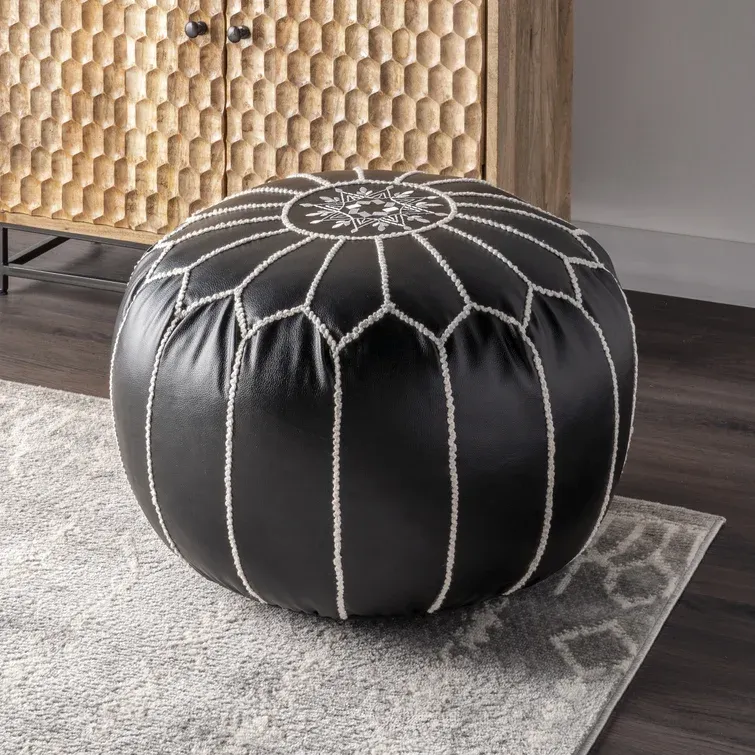 A black leather pouf with white stitching and a decorative design on top, placed on a light gray rug.