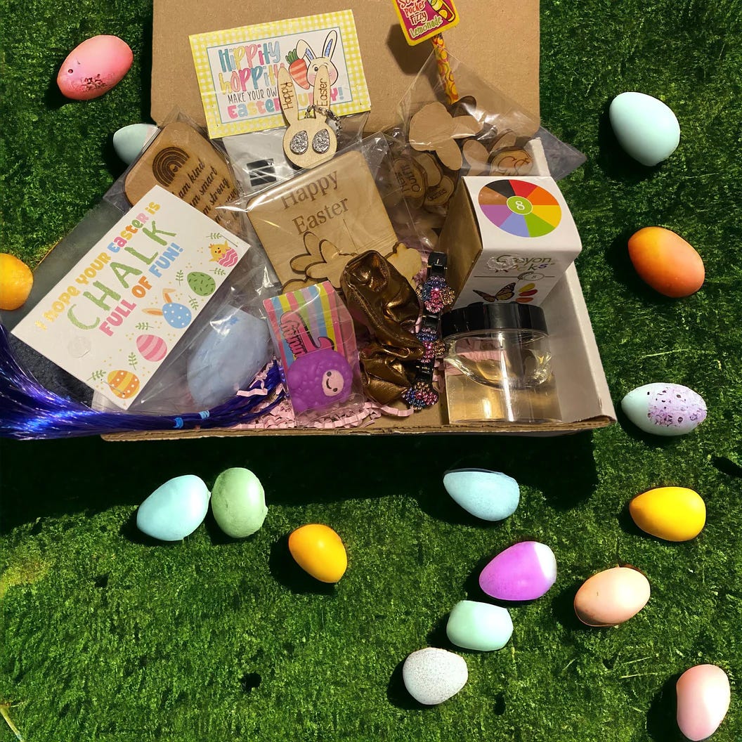 A box with Easter-themed items including chalk, decorations, and chocolate, surrounded by colorful eggs on grass.