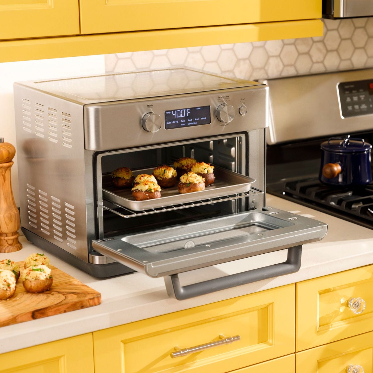 Stainless steel countertop oven with digital display cooking food, placed on a yellow kitchen counter beside a stovetop.