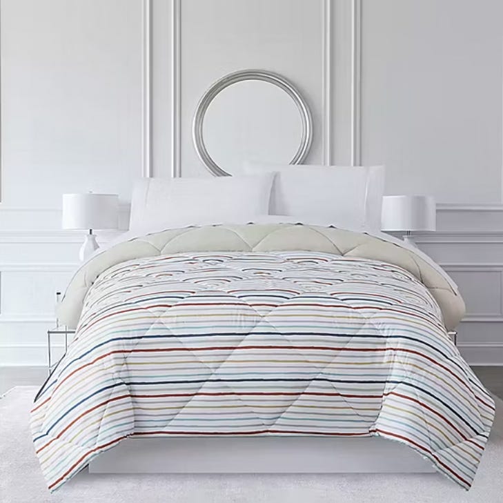 A neatly made bed features a white and multi-colored striped comforter, white pillows, and two bedside tables with white lamps. A circular mirror hangs on the wall above the bed.
