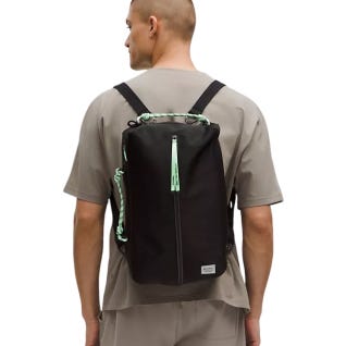 A man wearing a lululemon 16L Paracord Backpack, featuring a sleek black design with mint green paracord details, adjustable straps, and a top zipper closure. It's worn against a neutral-toned outfit.