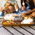 Indoor s'mores maker set with marshmallows, chocolate, graham crackers, pretzels, and strawberries on a wooden tray, surrounded by children.