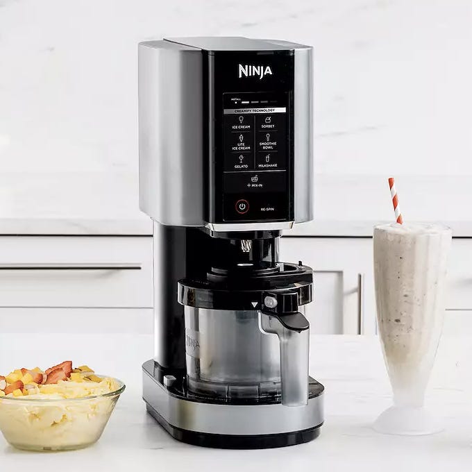 Ninja ice cream maker on a kitchen countertop next to a bowl of fruit and a milkshake glass with a straw.
