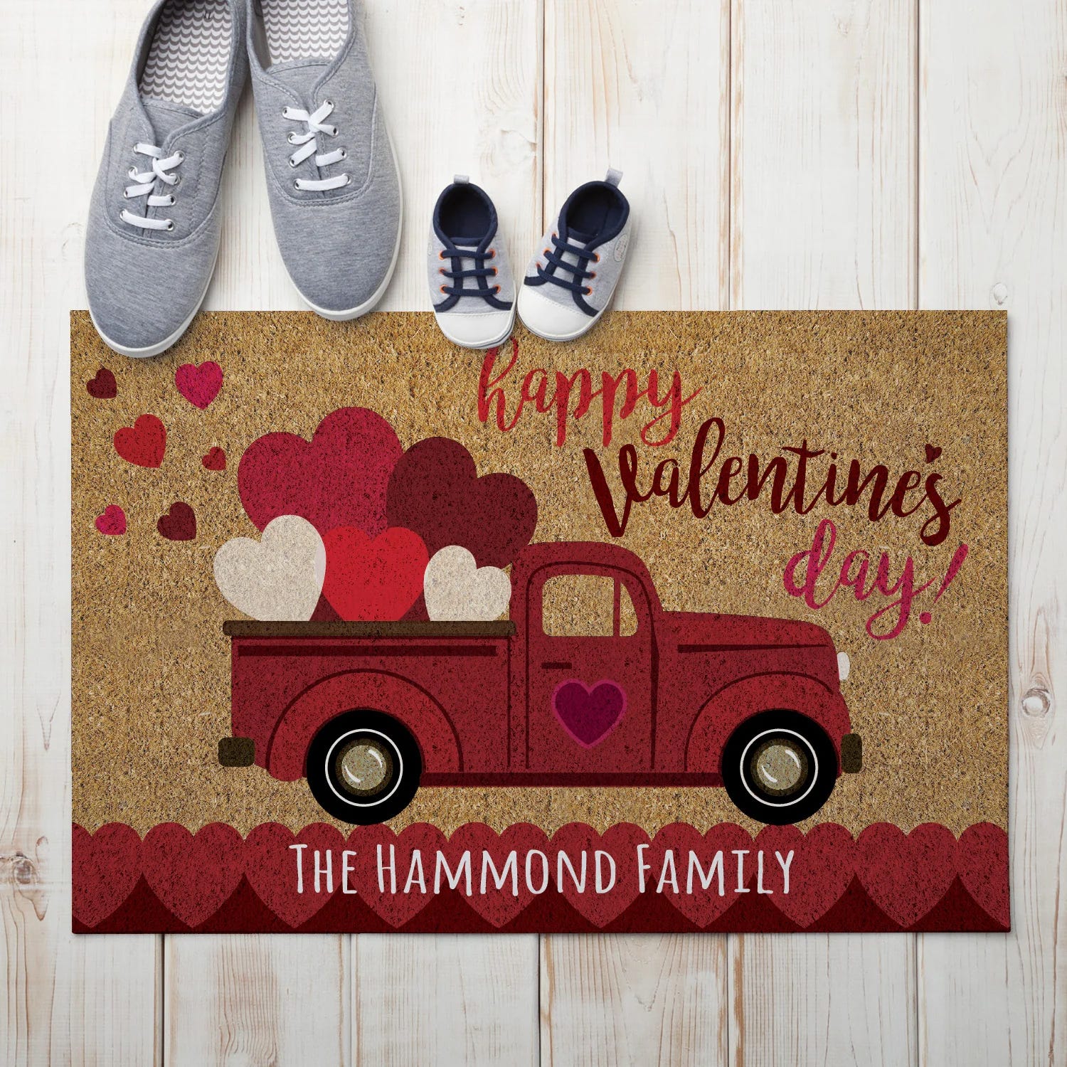 A doormat with a Valentine's Day theme features a red truck filled with hearts and the text \“Happy Valentine's Day!\“ and \“The Hammond Family.\“ Two pairs of shoes, one adult and one child-sized, are placed on a wooden floor beside it.