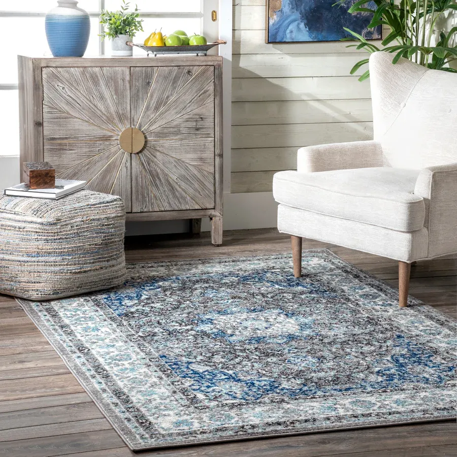 A weathered wooden cabinet, blue and beige patterned area rug, white fabric armchair with a footstool, and a woven pouf with a book on top.
