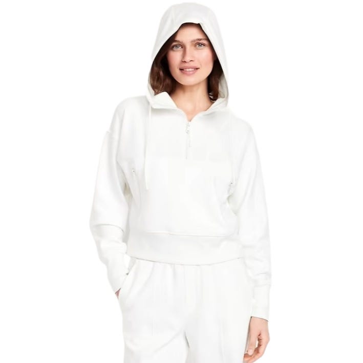 A woman is wearing a white hooded half-zip pullover sweatshirt paired with matching white pants, featuring a relaxed fit and drawstrings.